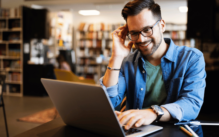 Smiling man wearing glasses working on a laptop at a desk.